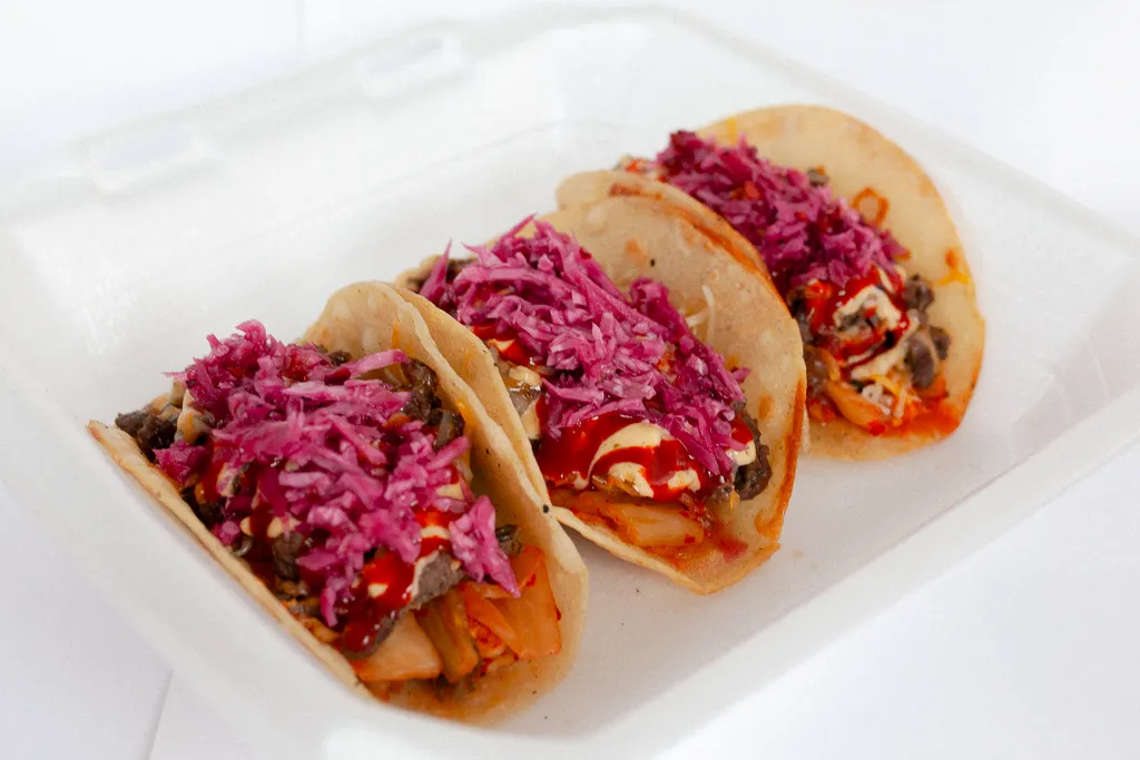 Three bulgogi tacos on corn tortillas topped with pickled red cabbage, kimchi, Seoul sauce and Sriracha